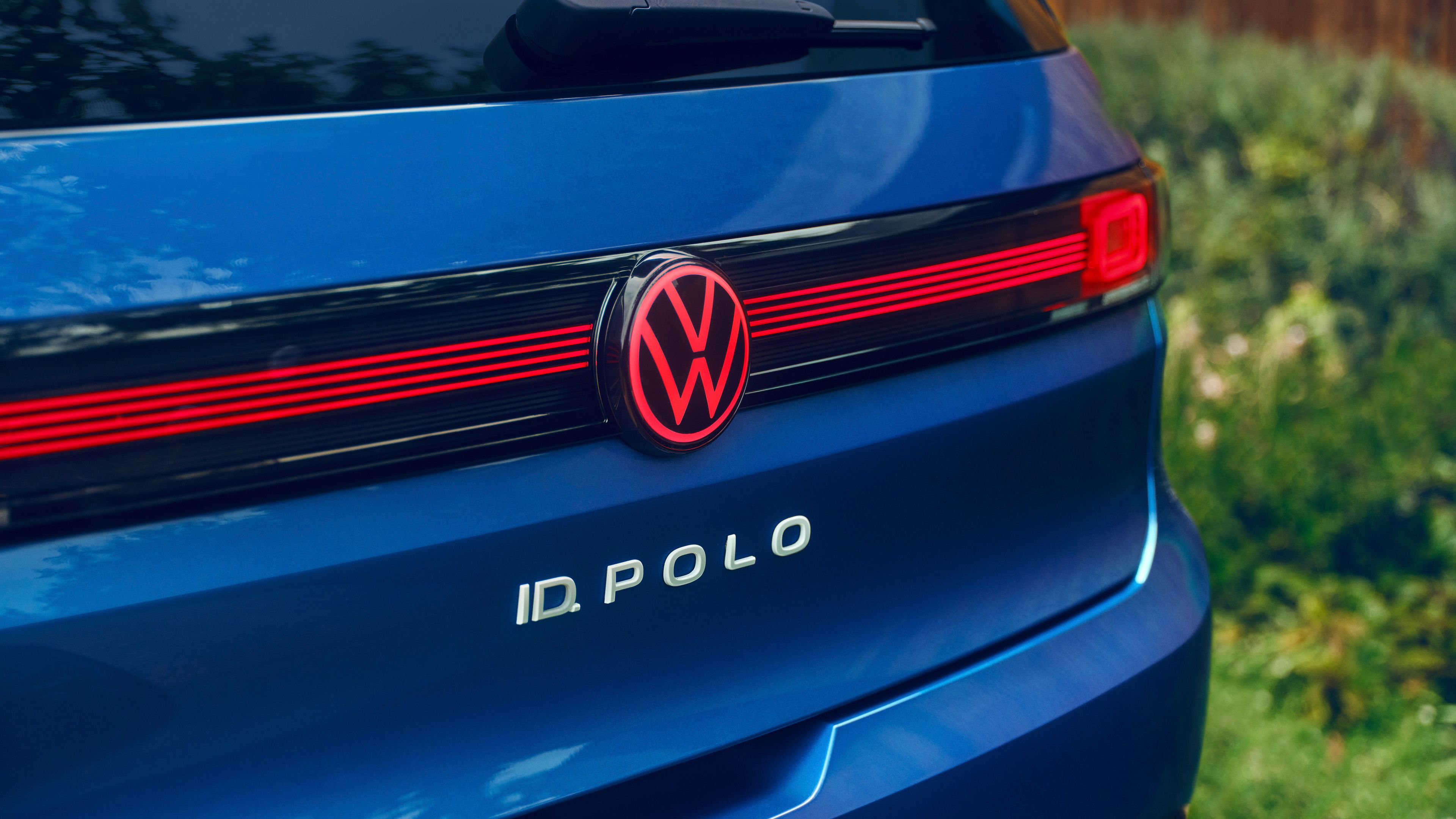 Zadnji del modrega ID. Pola z osvetljenim rdečim logotipom VW in tanko rdečo svetlobno letvijo čez celotno širino. Pod logotipom je bel napis modela “ID. POLO”. 
