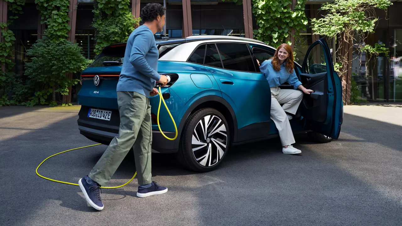 Moški z rumenim polnilnim kablom za električna vozila se približuje zadku parkiranega VW ID.4 v modri barvi. Skozi sovoznikova vrata iz vozila izstopa mlajša ženska.
