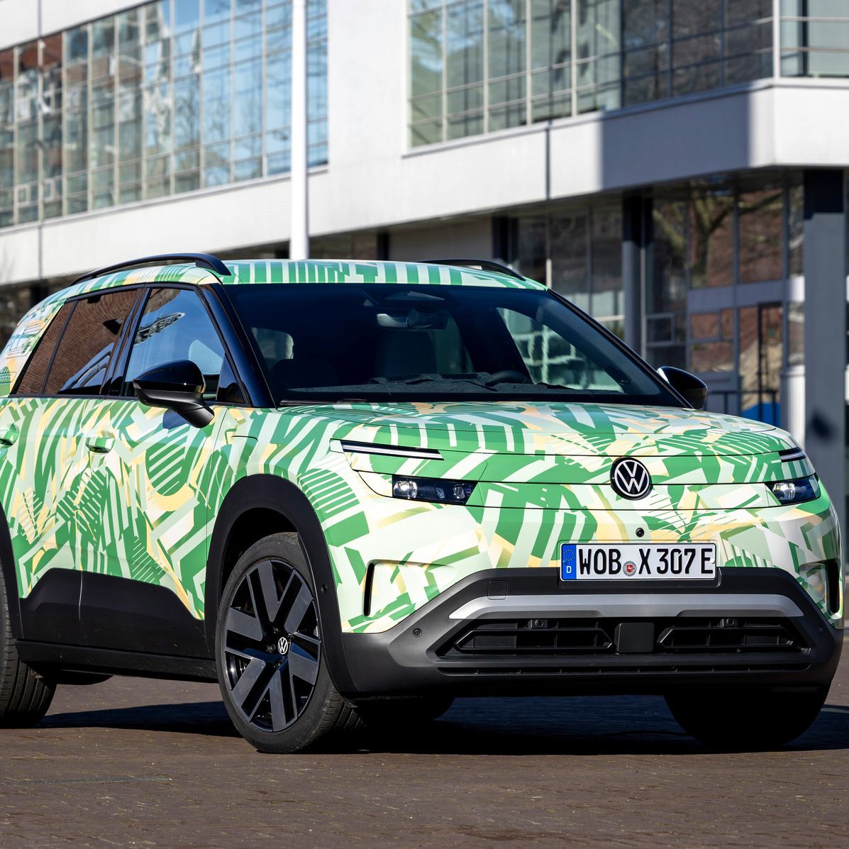 Pred sodobnimi stavbami stoji električni SUV Volkswagen. Barvito vozilo ima zeleno-kremno kamuflažno vzorec z motivi listov. 