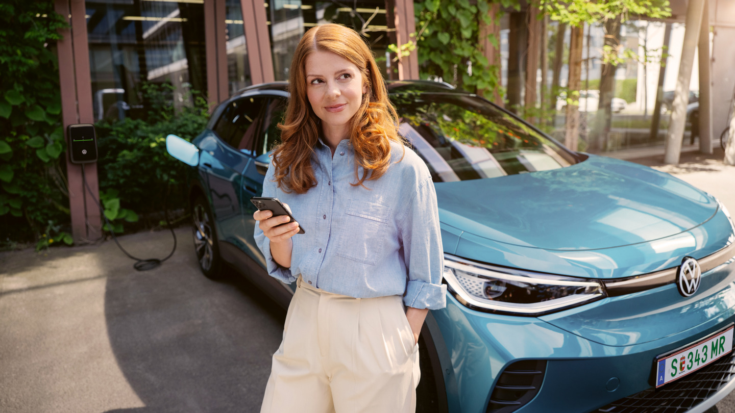 Ženska s pametnim telefonom v roki stoji pred modrim VW ID.4. V ozadju je vidna stenska polnilna postaja, na katero je priključeno vozilo.