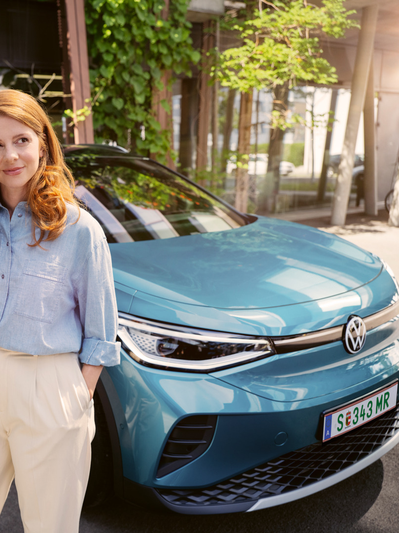 Ženska s pametnim telefonom v roki stoji pred modrim VW ID.4. V ozadju je vidna stenska polnilna postaja, na katero je priključeno vozilo.