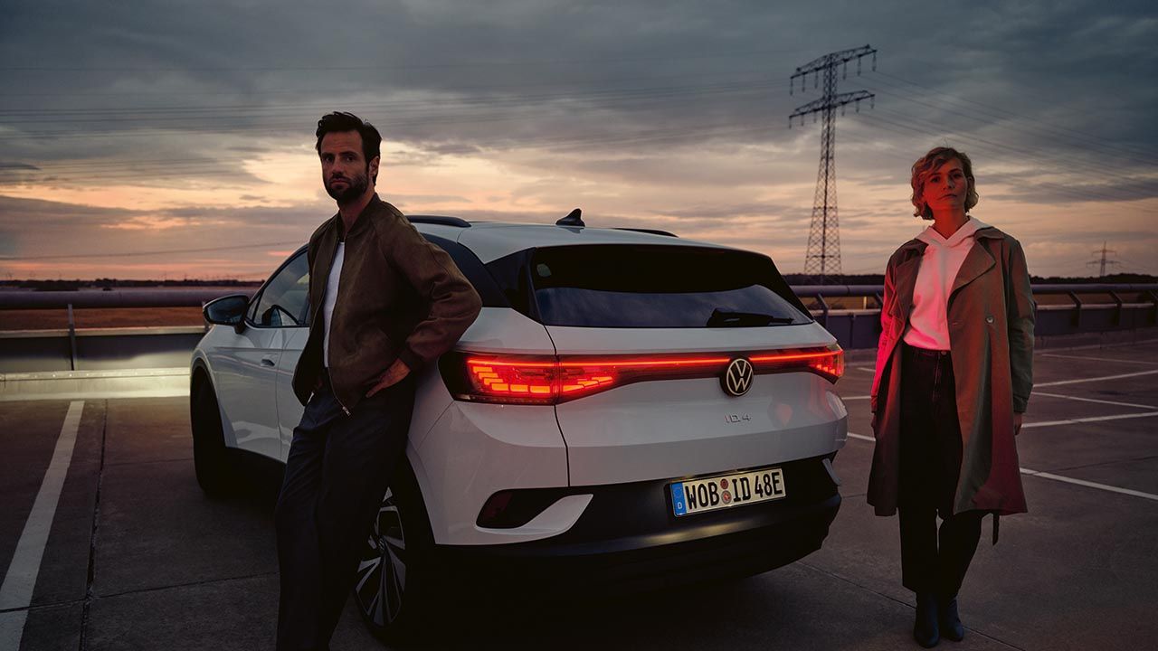 Bel VW ID.4 v mraku stoji na parkirišču. Zadnje luči osvetljujejo par za vozilom.