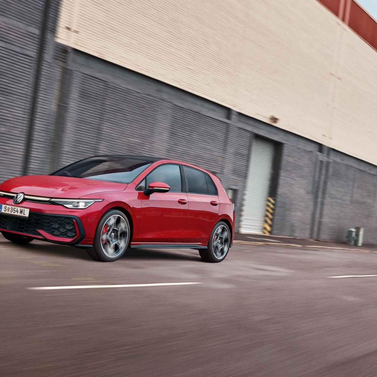 Diagonalno od spredaj je prikazan rdeč VW Golf GTI, ki se pelje mimo sive stavbe.