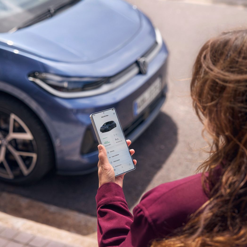 Ženska pogleda na svoj pametni telefon, pred njo je parkiran moder VW ID.3 Neo