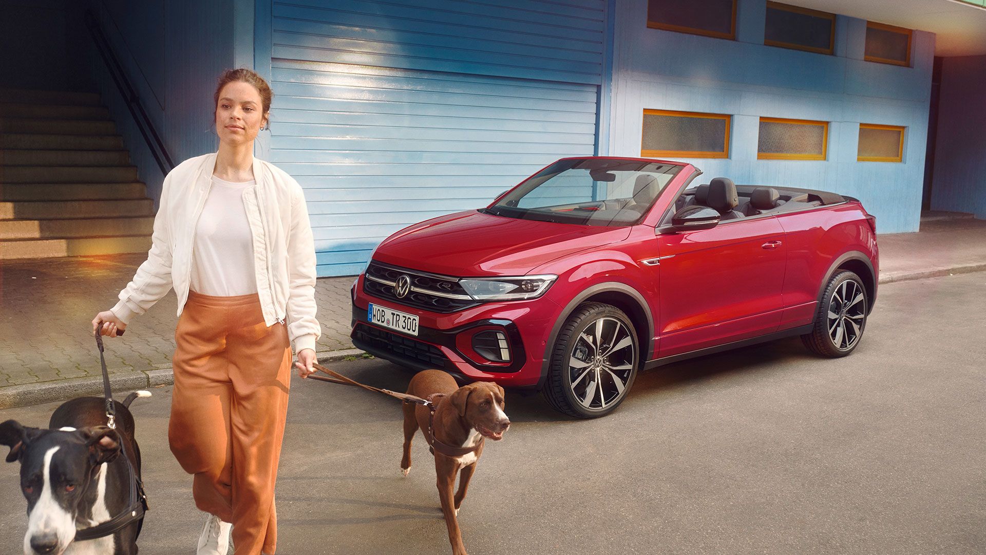 Rdeč VW T-Roc Cabriolet z odprto streho je parkiran ob robu ceste, v ospredju je ženska, ki z dvema psoma prečka cesto.
