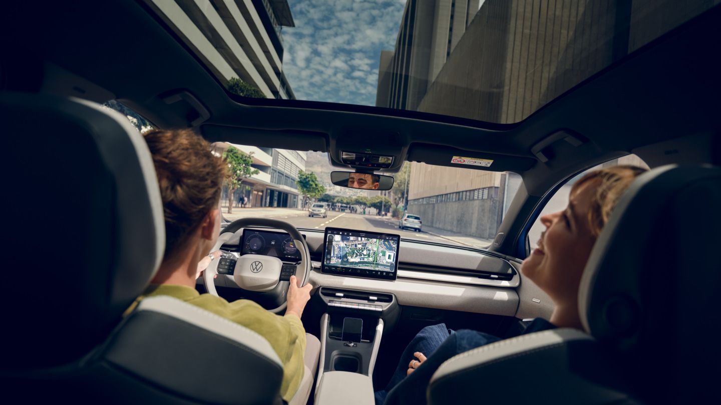 Moški vozi VW ID. Polo Style, na sovoznikovem sedežu ženska, ki se smeji. Visoke stavbe so vidne skozi panoramsko streho