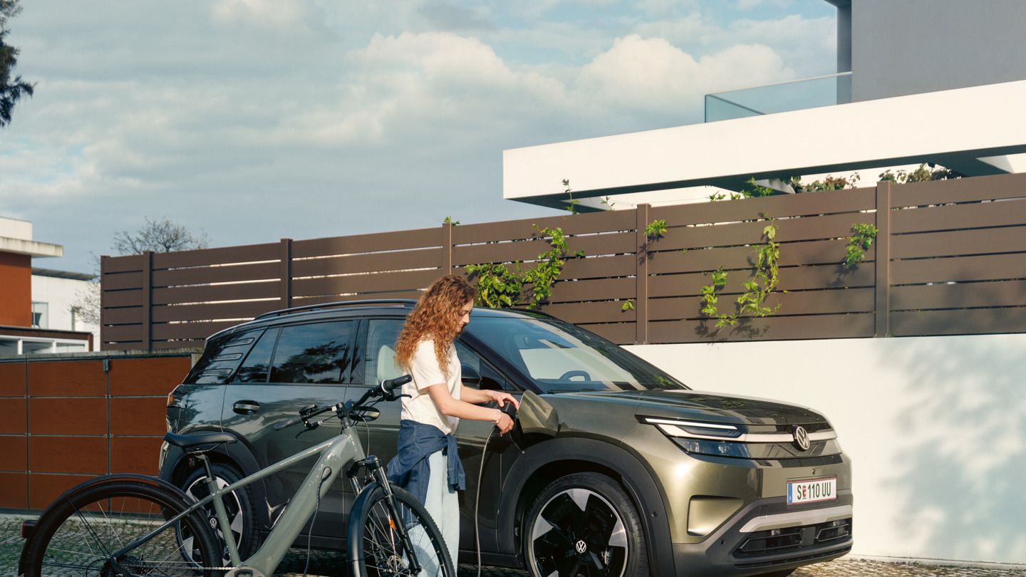 Ženska prek kabla polni električno kolo ob zelenem VW ID. Crossu, v ozadju je stanovanjska hiša.