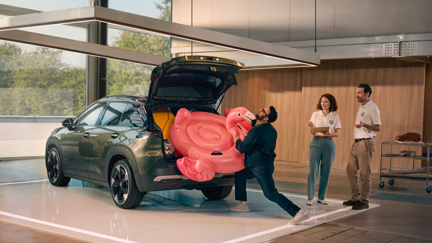 Moški potiska napihljivega flaminga v odprt prtljažnik VW ID. Crossa. Ob njem stojita dve osebi in opazujeta.