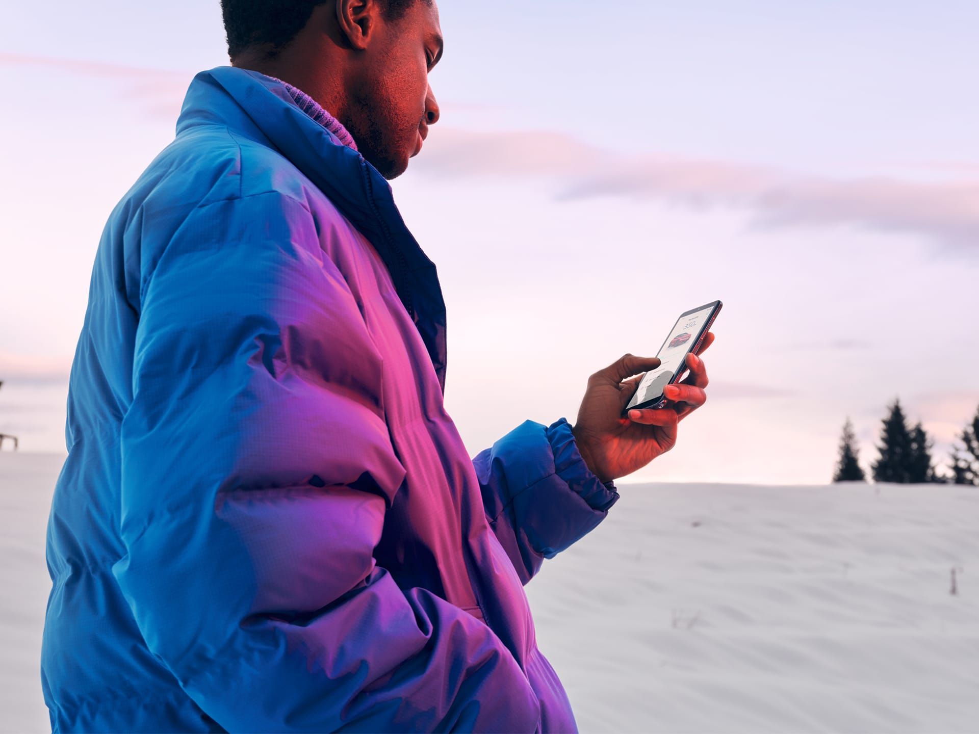 Ein Mann in Winterjacke steht in verschneiter Landschaft und schaut auf sein Smartphone.