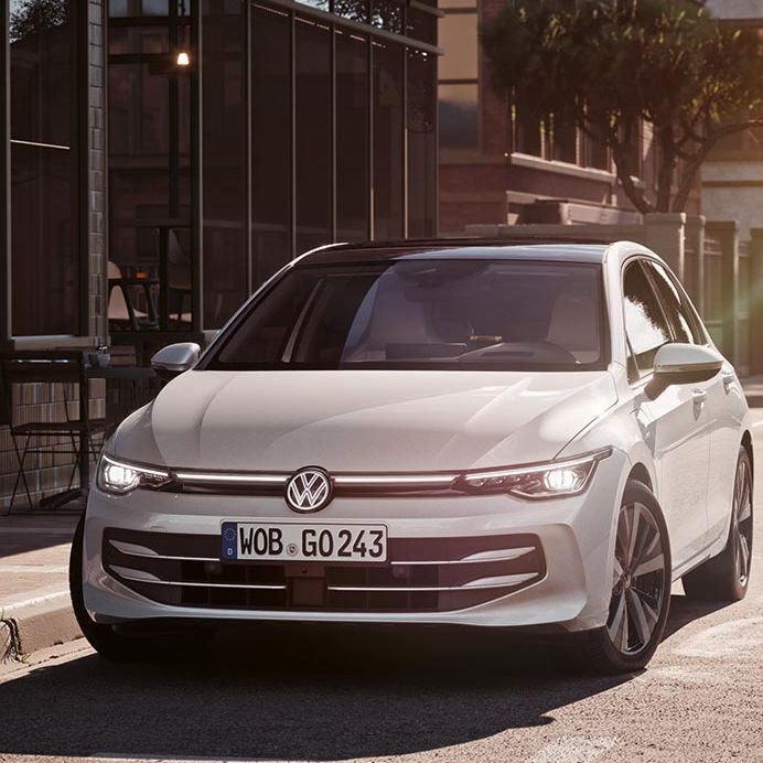 Bel avtomobil Volkswagen Golf stoji ob robu sončne mestne ulice, obrnjen rahlo proti kameri. Stavbe z veliko stekla in drevesa v ozadju nakazujejo mirno urbano okolje. Svetloba poznega popoldneva ustvarja mehke sence na cesti in karoseriji.