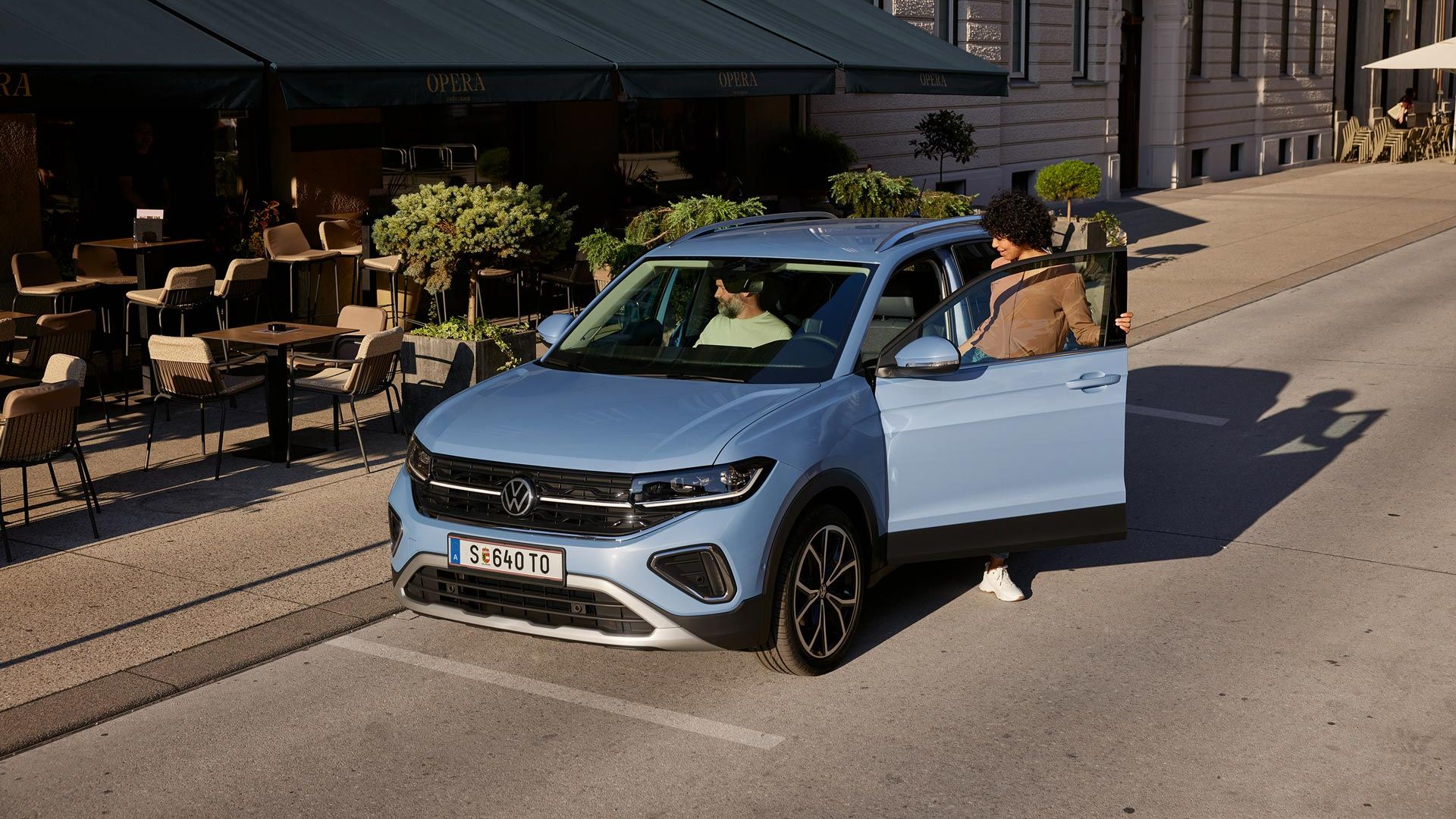 Ženska na voznikovi strani vstopa v moder VW T-Cross, ki je parkiran pred mestno ulično kavarno.