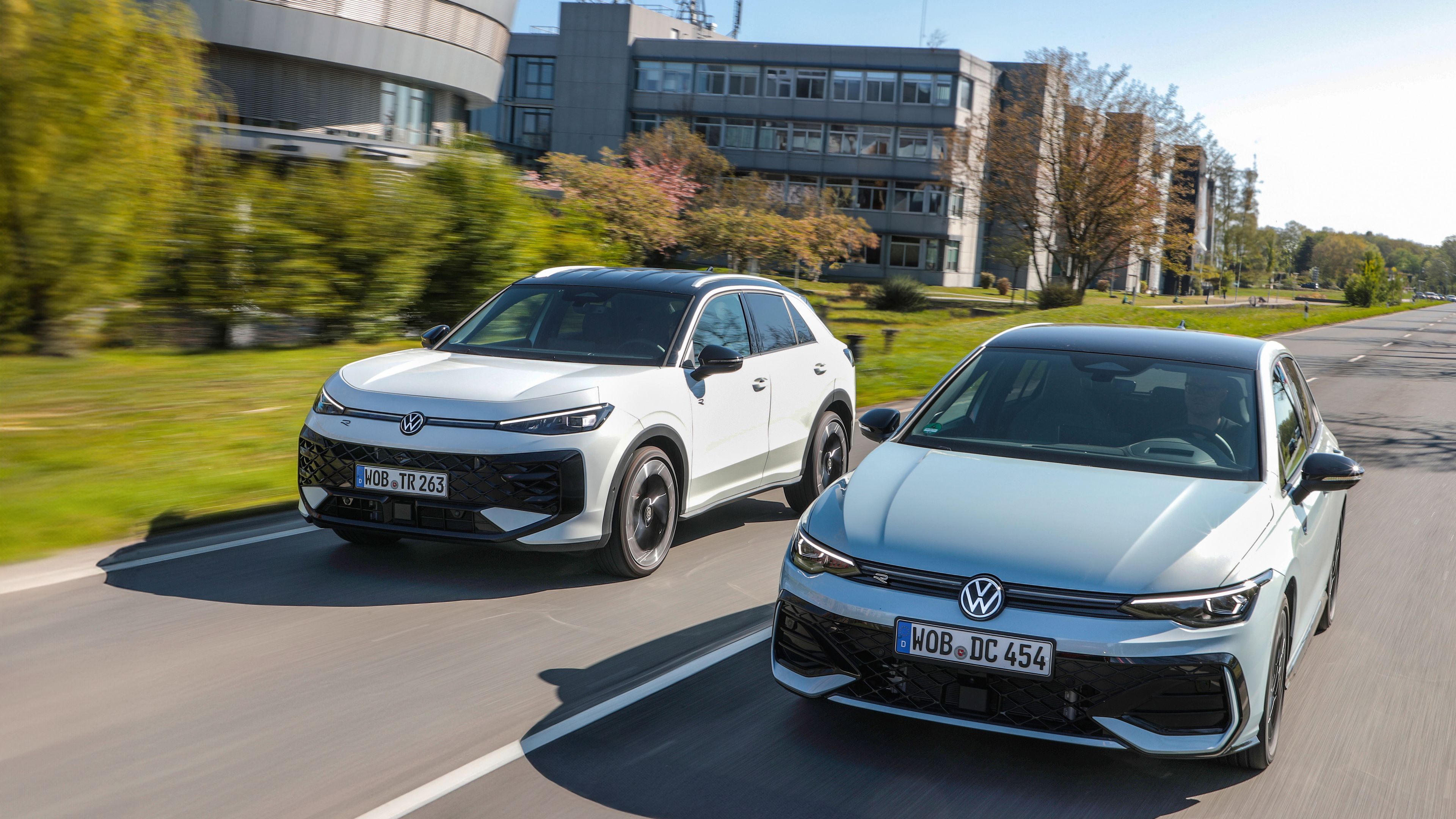 Volkswagen T-Roc SUV in Golf se vozita vzporedno po dvopasovni cesti. V ozadju so poslovne zgradbe, drevesa in trava, kar nakazuje mestno ali predmestno okolje. Obe vozili sta v gibanju, posneti iz rahlega sprednjega kota.