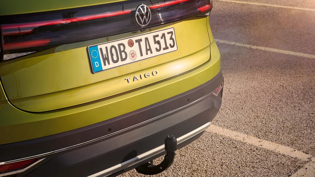 Zadnji del svetlo zelenega Volkswagna Taiga, parkiranega na asfaltiranem parkirišču. V ospredju je vidna vlečna kljuka, nad njo pa registrska tablica z evropsko oznako in napisom WOB TA 513. Na zadku vozila poteka vodoravna LED zadnja luč.