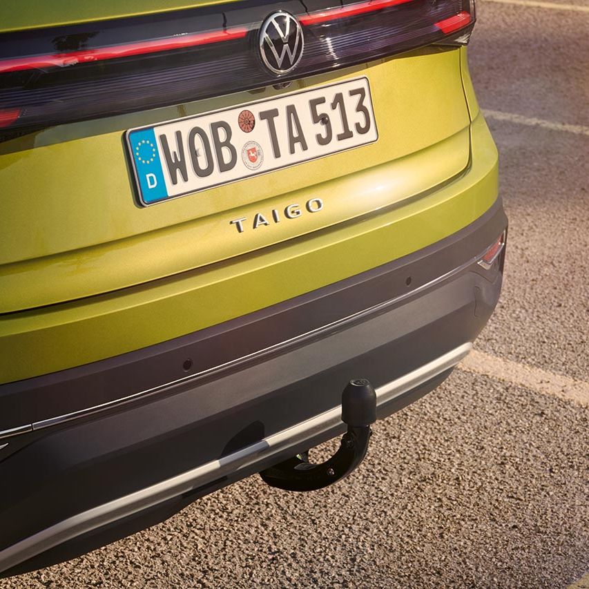 Zadnji del svetlo zelenega Volkswagna Taiga, parkiranega na asfaltiranem parkirišču. V ospredju je vidna vlečna kljuka, nad njo pa registrska tablica z evropsko oznako in napisom WOB TA 513. Na zadku vozila poteka vodoravna LED zadnja luč.