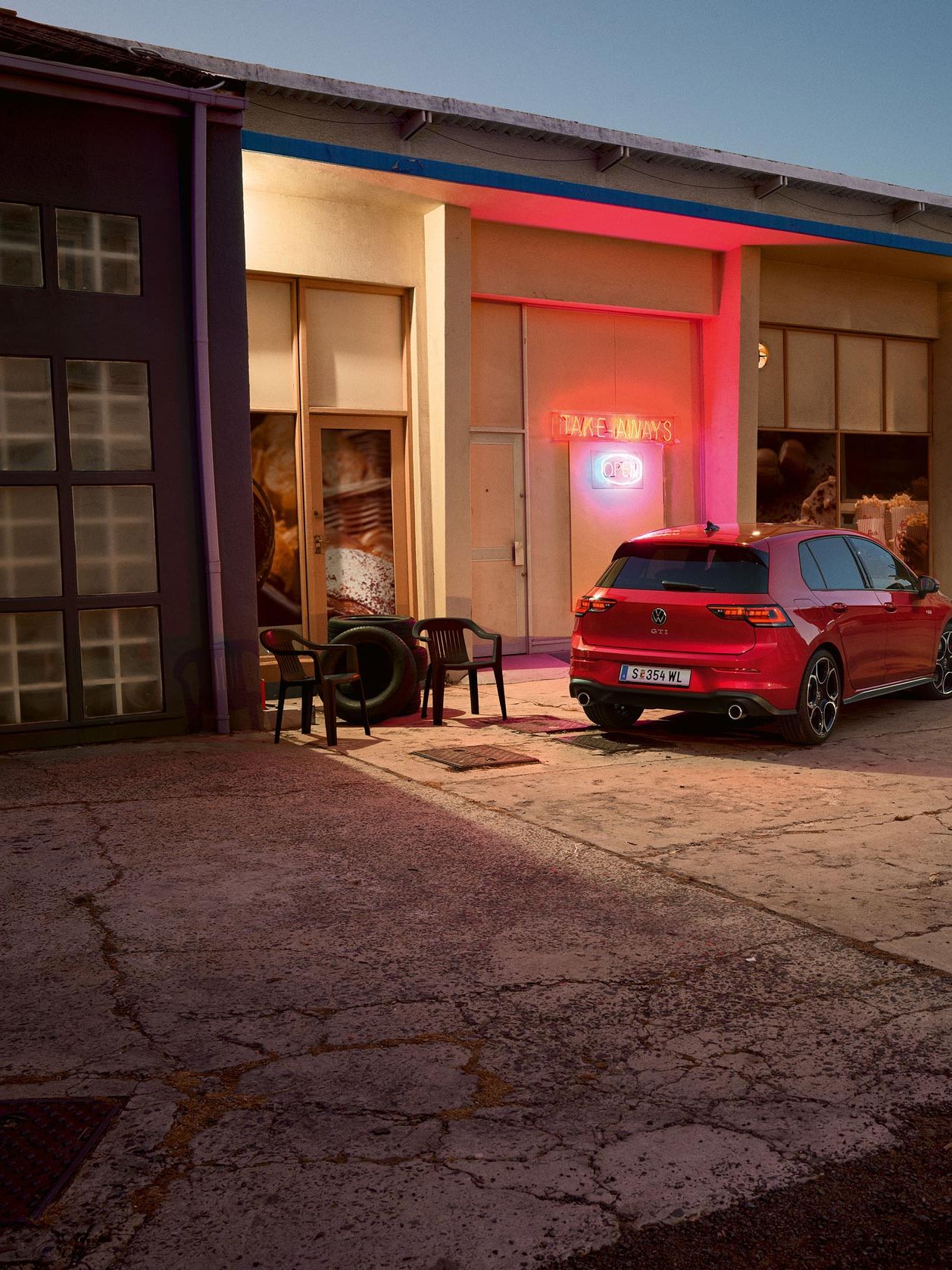 Zadek rdečega VW Golfa GTI, ki je parkiran pred kioskom. Moški in ženska s prigrizki v rokah se približujeta.