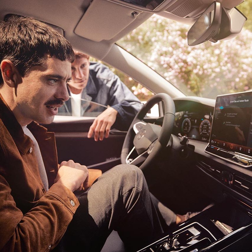 Moški sedi na voznikovem sedežu VW Golf Variant, ob odprtem oknu se z njim pogovarja druga oseba, naslonjena na vrata. Na sredinski konzoli je velik zaslon na dotik z vklopljeno navigacijo in multimedijo, notranjost vozila je svetla in urejena. V ozadju se skozi okna vidijo drevesa in hiše v sončnem vremenu.