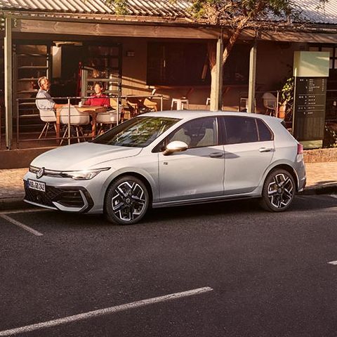 Ulični prizor z majhno restavracijo ali kavarno z zunanjimi mizami pod nadstreškom na levi strani slike. Pred lokalom je na cesti parkiran srebrn Volkswagen Golf.