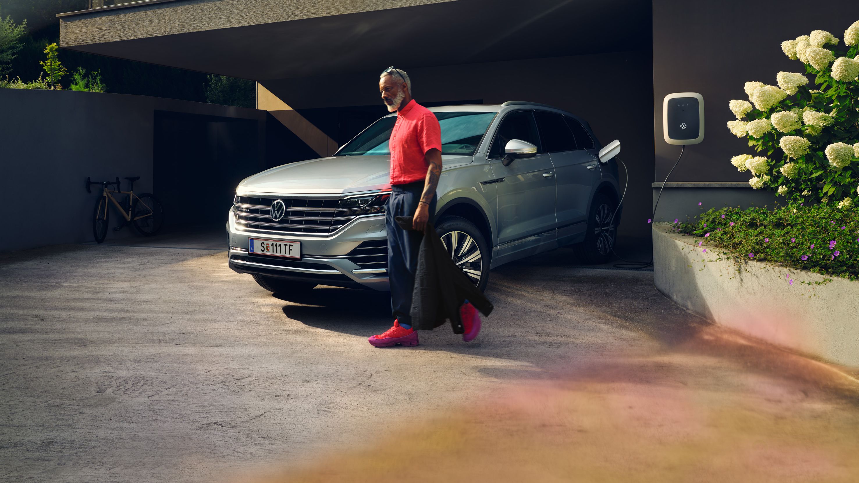 Volkswagen Touareg eHybrid srebrne barve, pogled na sprednji in stranski del, polni se na dovozu, moški gre mimo