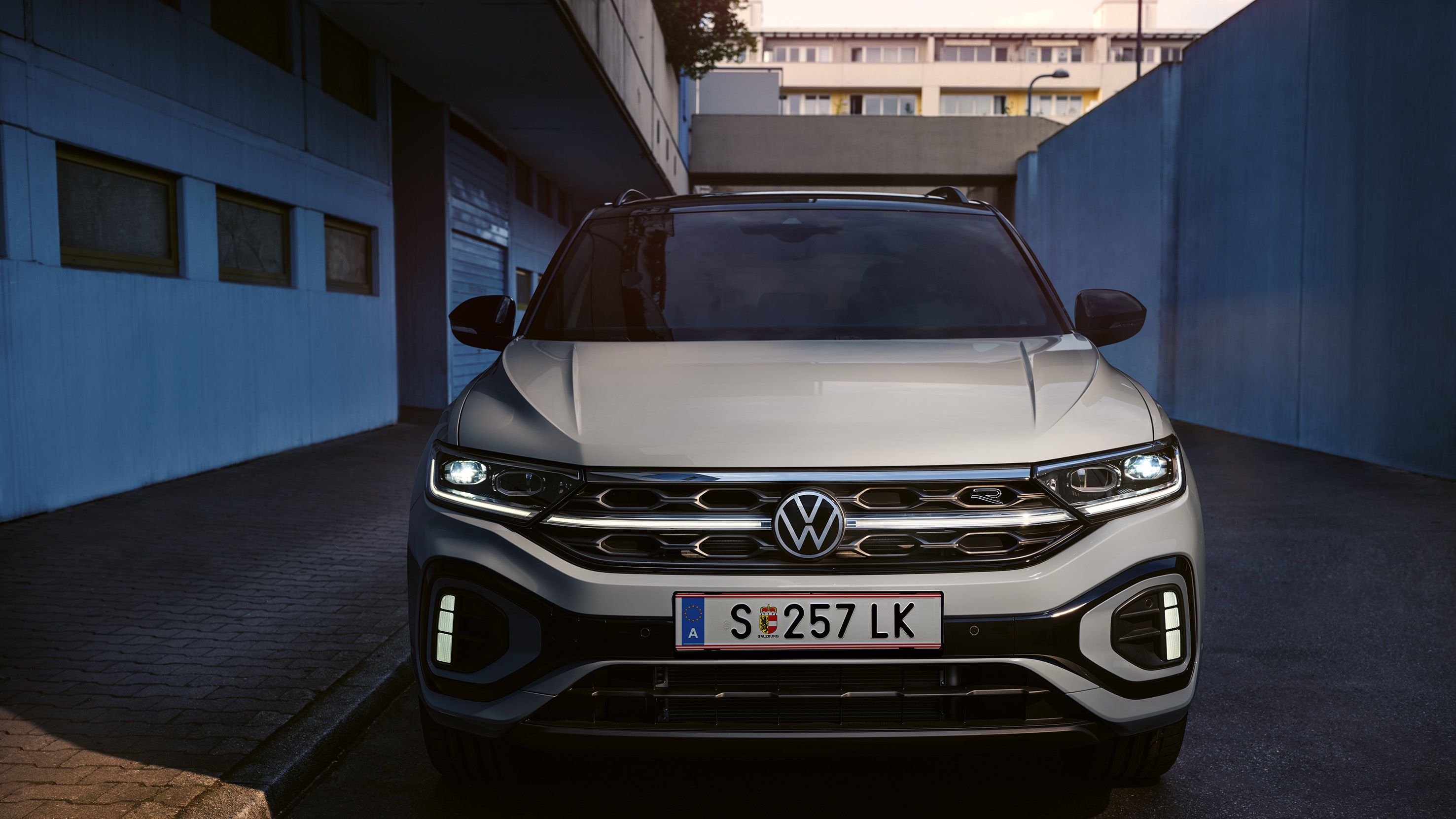 Sprednji del belega VW T-Roca R-Line, svetijo dnevne LED-luči in opcijska, osvetljena mreža hladilnika.
