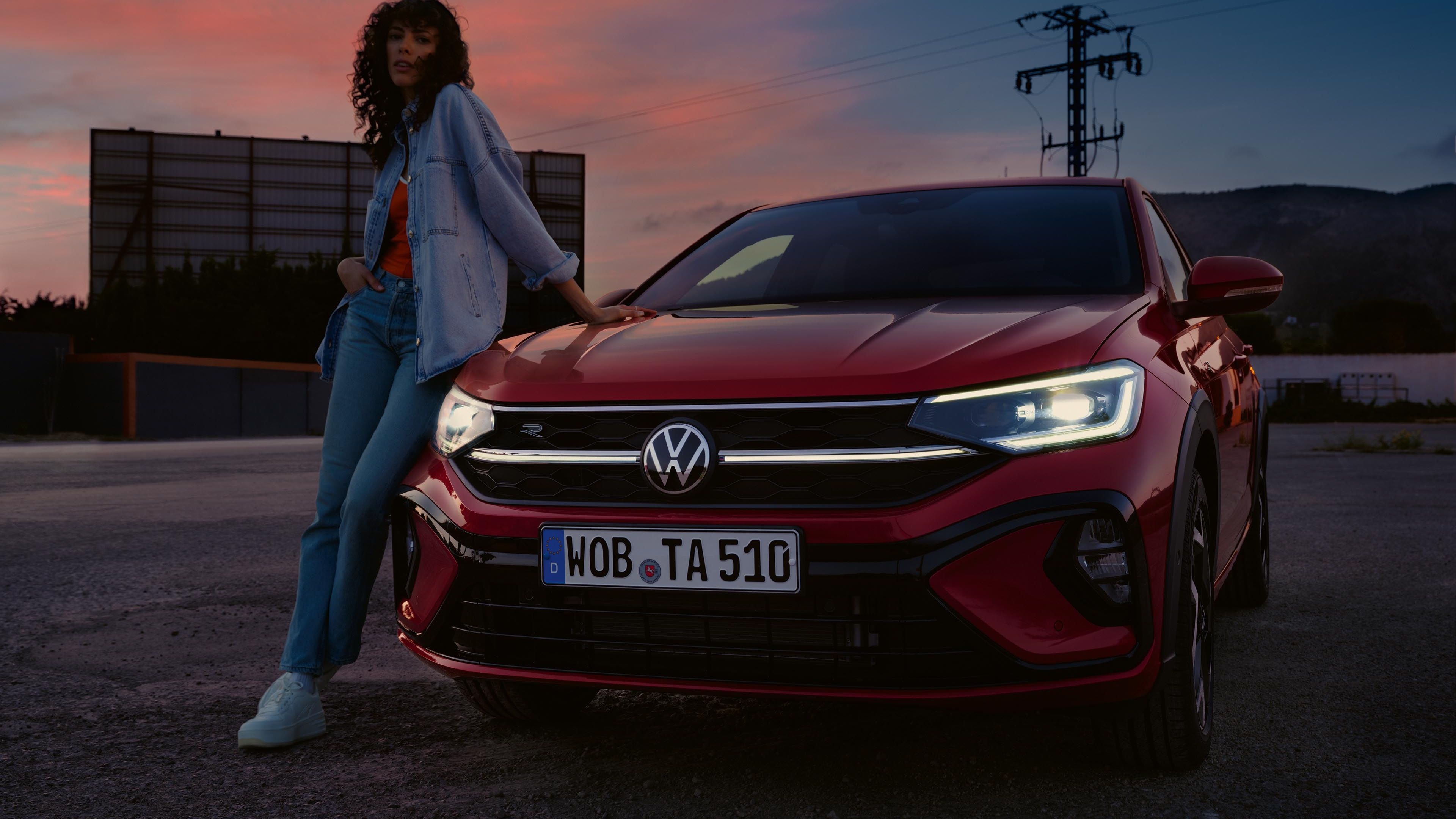 Rdeč VW Taigo R-Line na parkirišču. Luči so vključene. Ženska se naslanja na pokrov motornega prostora.