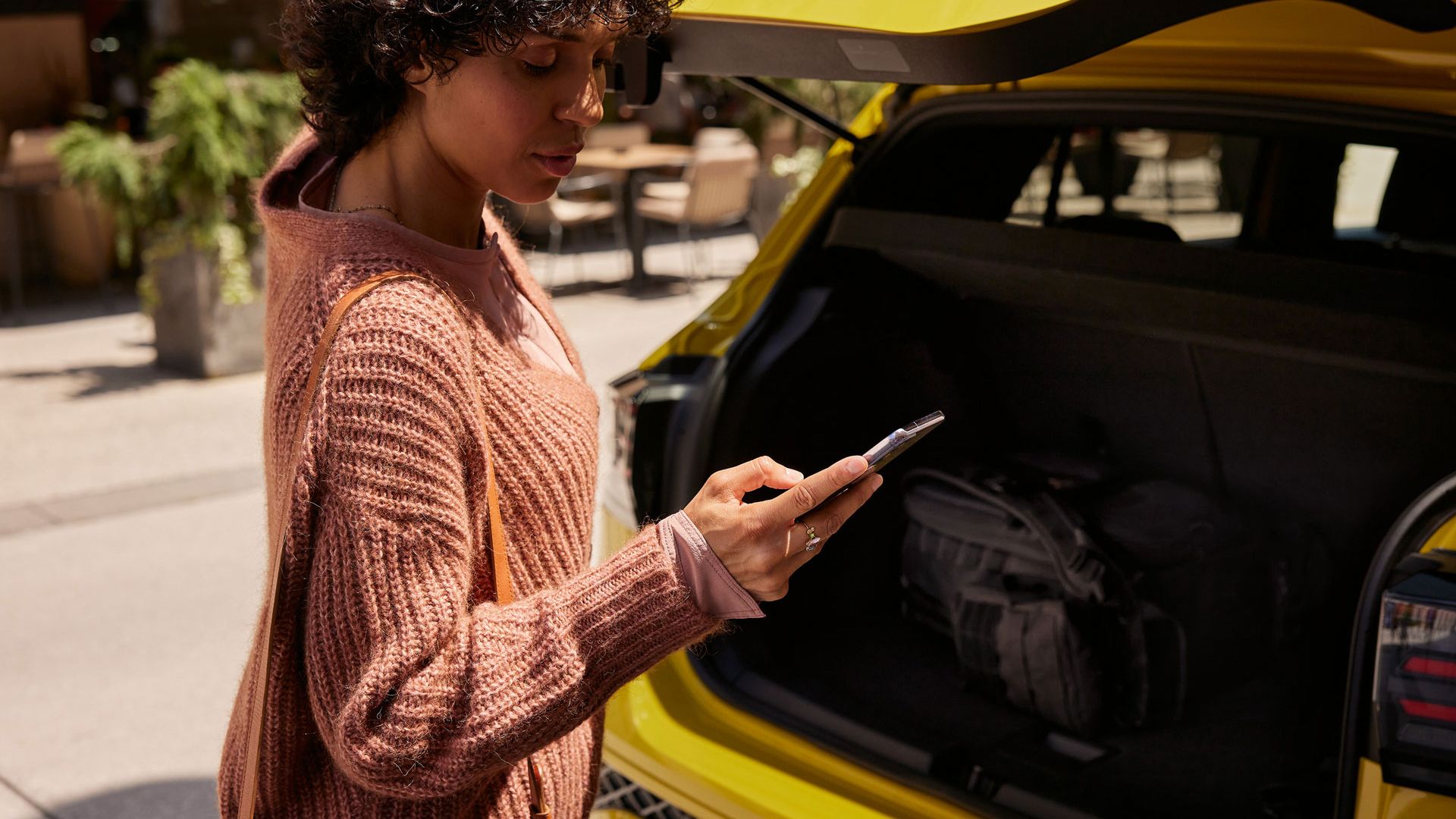 Eine frau schließt den Kofferraum eines gelben VW T-Cross und blickt dabei auf ihr Smartphone. 