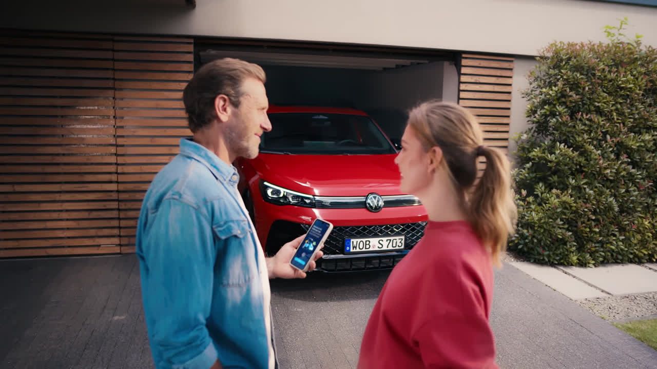 Moški in ženska stojita pred sodobno hišo, moški v roki drži pametni telefon z odprto aplikacijo. Iz garaže za njima vzvratno zapelje rdeč Volkswagnov avto. Hiša ima lesene panele, ob strani pa je grm zelenja.