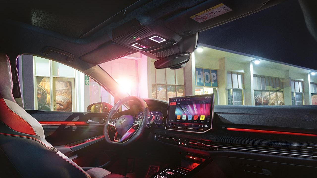 Notranjost Volkswagen Golf GTI-ja ponoči z osvetljenim ambientom v rdečih odtenkih. Na sredinski konzoli je velik zaslon na dotik z multimedijskim vmesnikom, obkrožajo pa ga elegantne, ravne linije armaturne plošče. skozi sprednja in stranska stekla se vidi zunanjost z osvetljeno poslovalnico ali bencinsko črpalko.