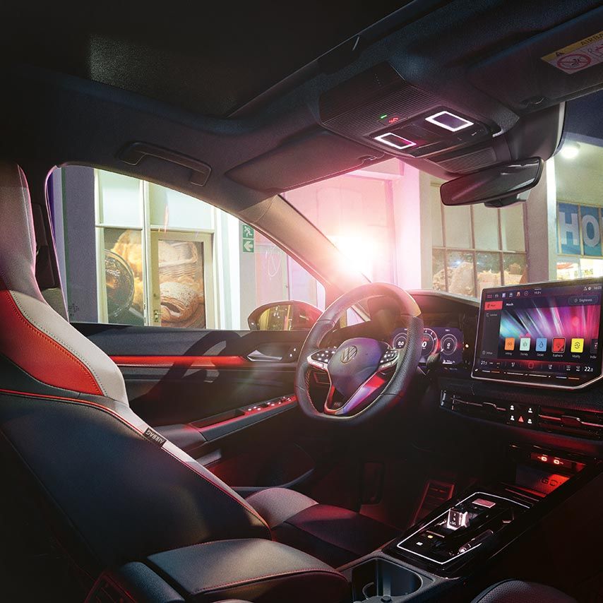 Notranjost Volkswagen Golf GTI-ja ponoči z osvetljenim ambientom v rdečih odtenkih. Na sredinski konzoli je velik zaslon na dotik z multimedijskim vmesnikom, obkrožajo pa ga elegantne, ravne linije armaturne plošče. skozi sprednja in stranska stekla se vidi zunanjost z osvetljeno poslovalnico ali bencinsko črpalko.