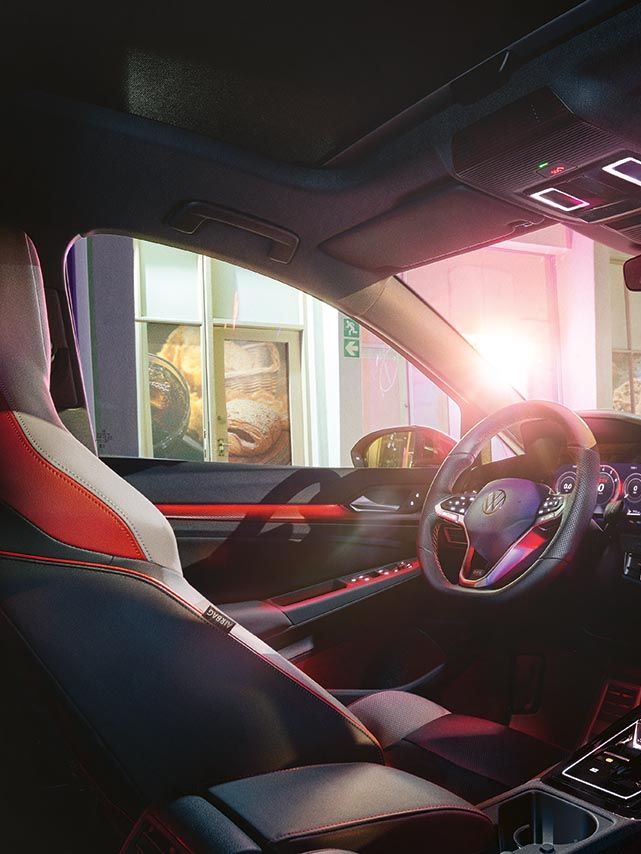Notranjost Volkswagen Golf GTI-ja ponoči z osvetljenim ambientom v rdečih odtenkih. Na sredinski konzoli je velik zaslon na dotik z multimedijskim vmesnikom, obkrožajo pa ga elegantne, ravne linije armaturne plošče. skozi sprednja in stranska stekla se vidi zunanjost z osvetljeno poslovalnico ali bencinsko črpalko.