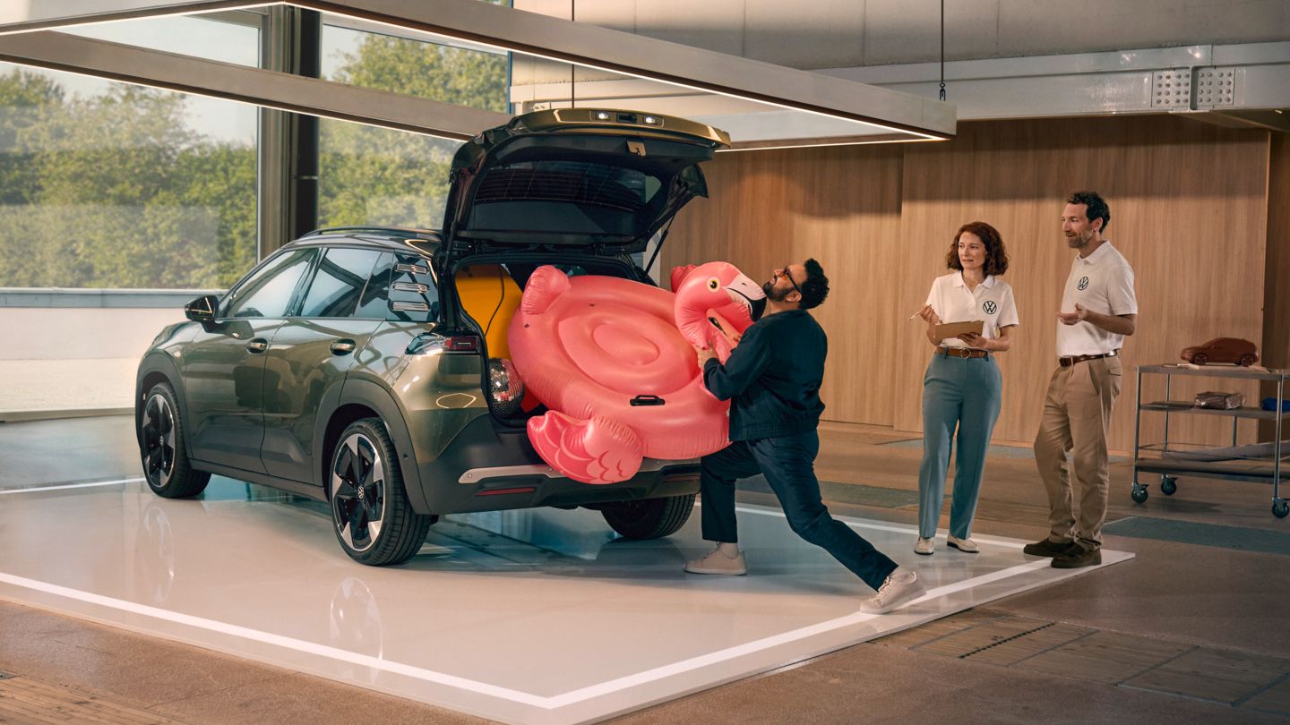 Moški potiska napihljivega flaminga v odprt prtljažnik VW ID. Crossa. Ob njem stojita dve osebi in opazujeta.