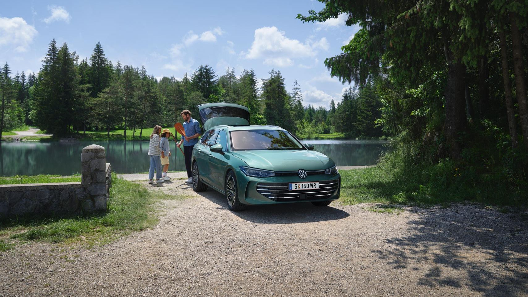 VW Passat in blaugrün parkt an einem Badesee mit geöffnetem Kofferraum. Ein Mann verteilt Freitzeitspielzeug an zwei Kinder.