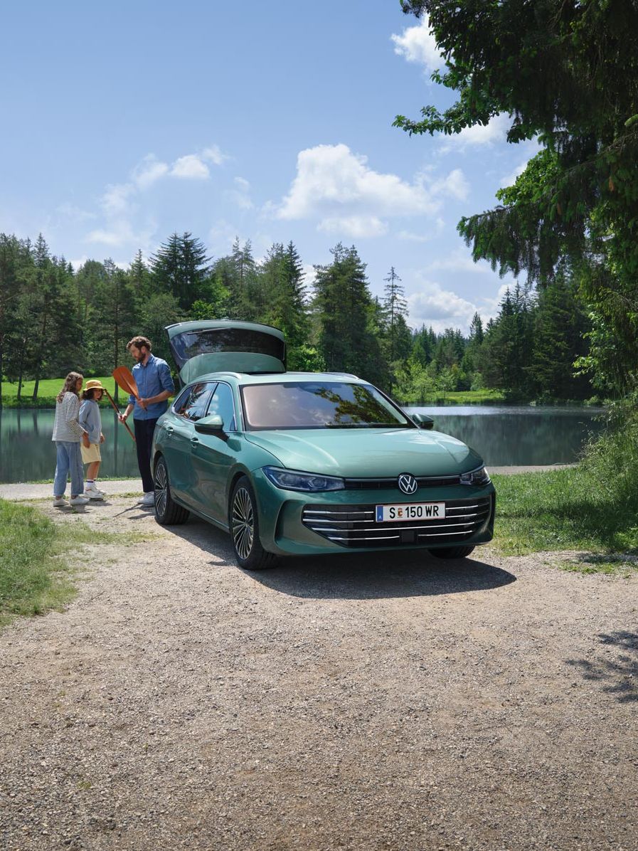 VW Passat in blaugrün parkt an einem Badesee mit geöffnetem Kofferraum. Ein Mann verteilt Freitzeitspielzeug an zwei Kinder.