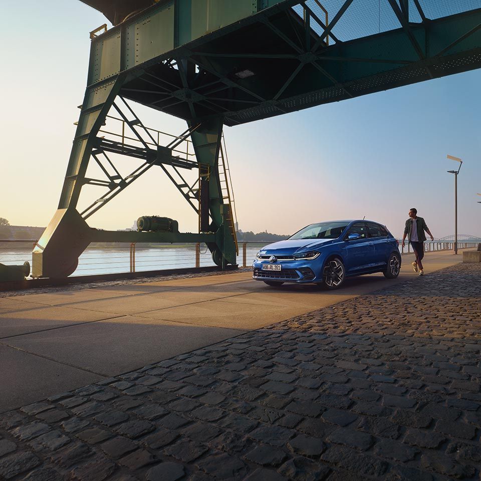 Moderen Volkswagen Polo modre barve je parkiran na tlakovanem nabrežju ob reki, pod masivno kovinsko konstrukcijo mostu ali dvigala. Ob avtomobilu hodi elegantno oblečen moški z aktovko v roki. V ozadju se ob reki razteza industrijsko-urbano okolje v mehki svetlobi sončnega zahoda.