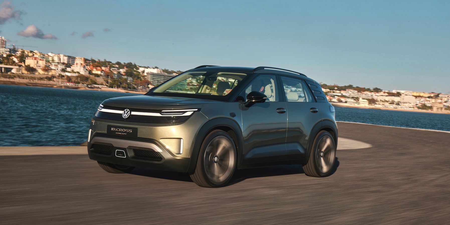 Zeleni Volkswagen ID. Cross vozi ob vodnem telesu ob obalni cesti. V ozadju se vidijo stavbe in jasno modro nebo. 