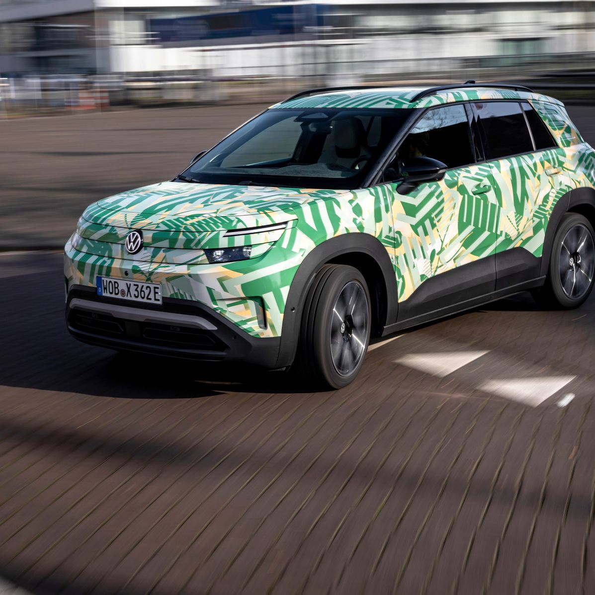 Avto z zeleno in rumeno kamuflažno barvo vozi po ulici. Gre za električni Volkswagen SUV . V ozadju so vidne zamegljene stavbe.