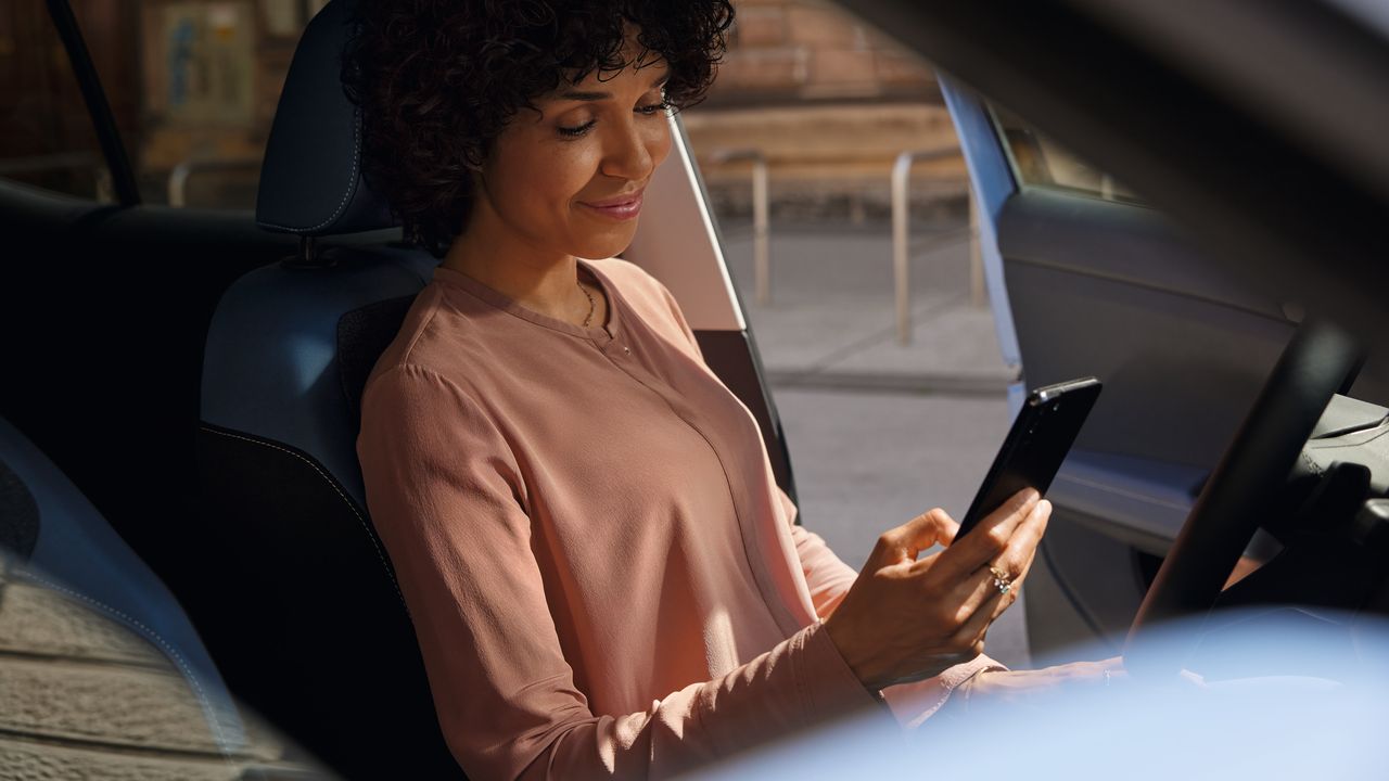 Nasmejana ženska sedi na voznikovem sedežu parkiranega VW T-Crossa in gleda v svoj mobilni telefon.