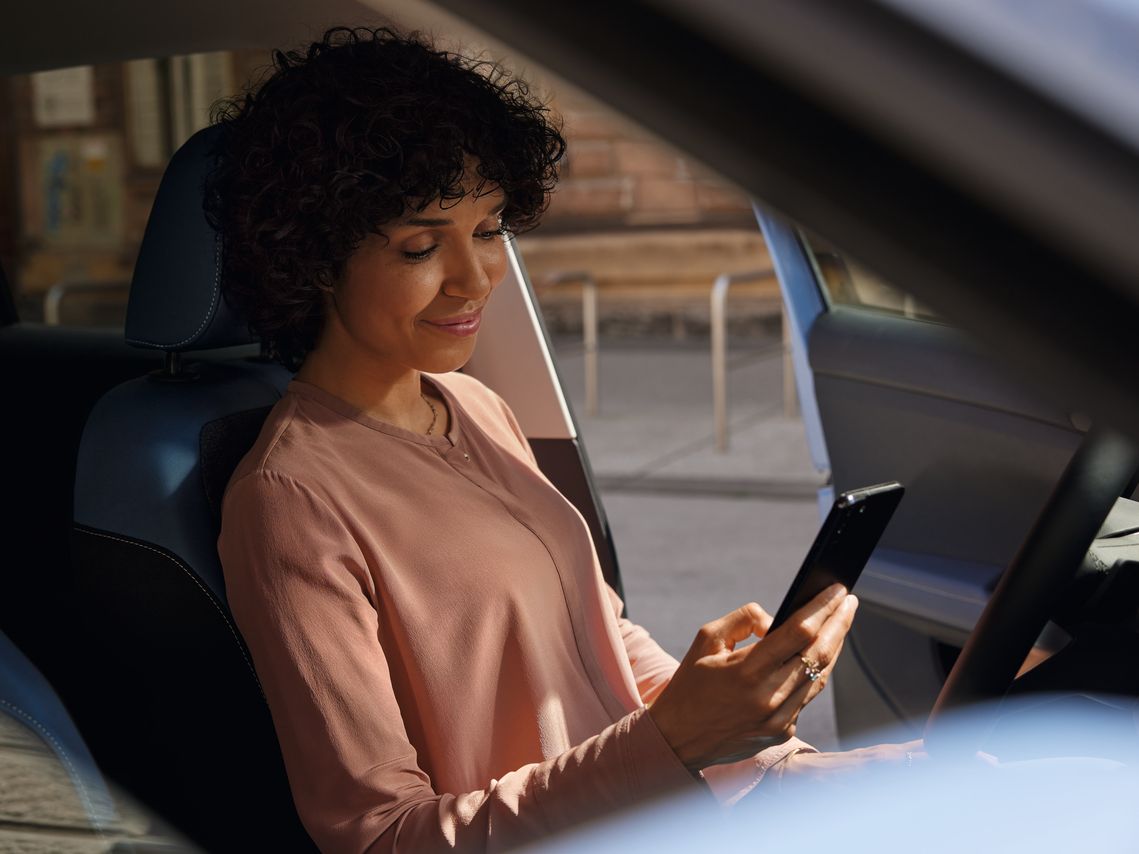 Nasmejana ženska sedi na voznikovem sedežu parkiranega VW T-Crossa in gleda v svoj mobilni telefon.