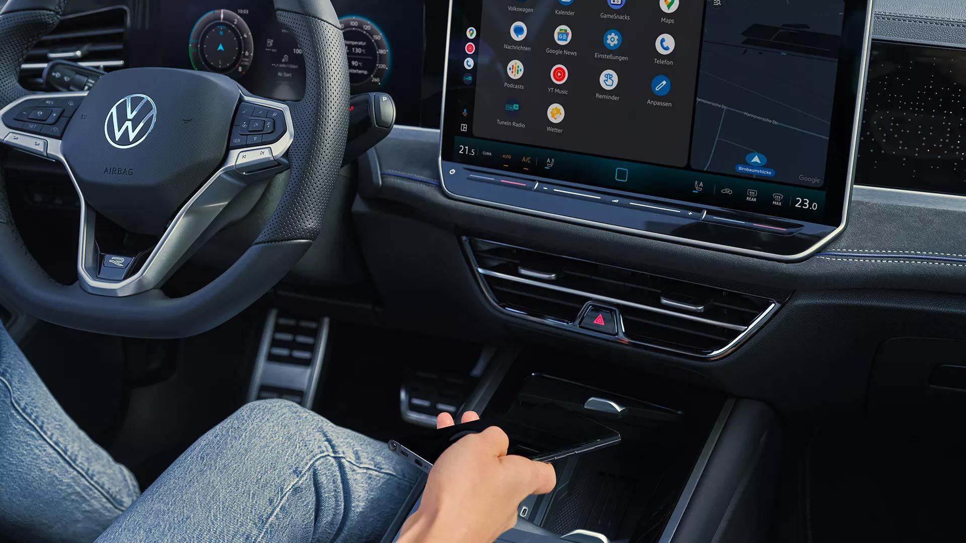 Detajlni posnetek armaturne plošče v VW Passatu. Oseba na voznikovem sedežu odlaga pametni telefon v predal v sredinski konzoli.
