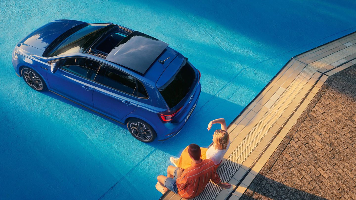 Moder VW Polo je parkiran v praznem bazenu, na robu bazena sedi parček. Od zgoraj je s strani prikazano opcijsko panoramsko dvižno-pomično strešno okno.