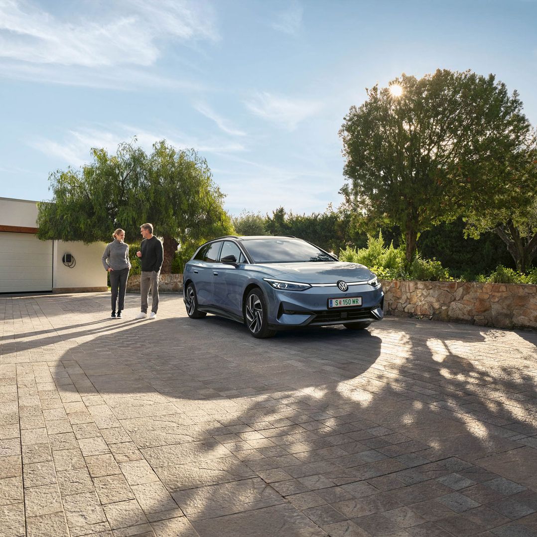 Der VW ID.7 Tourer ist vor Einfamillienhaus geparkt, eine Frau und ein Mann stehen neben ihm und unterhalten sich