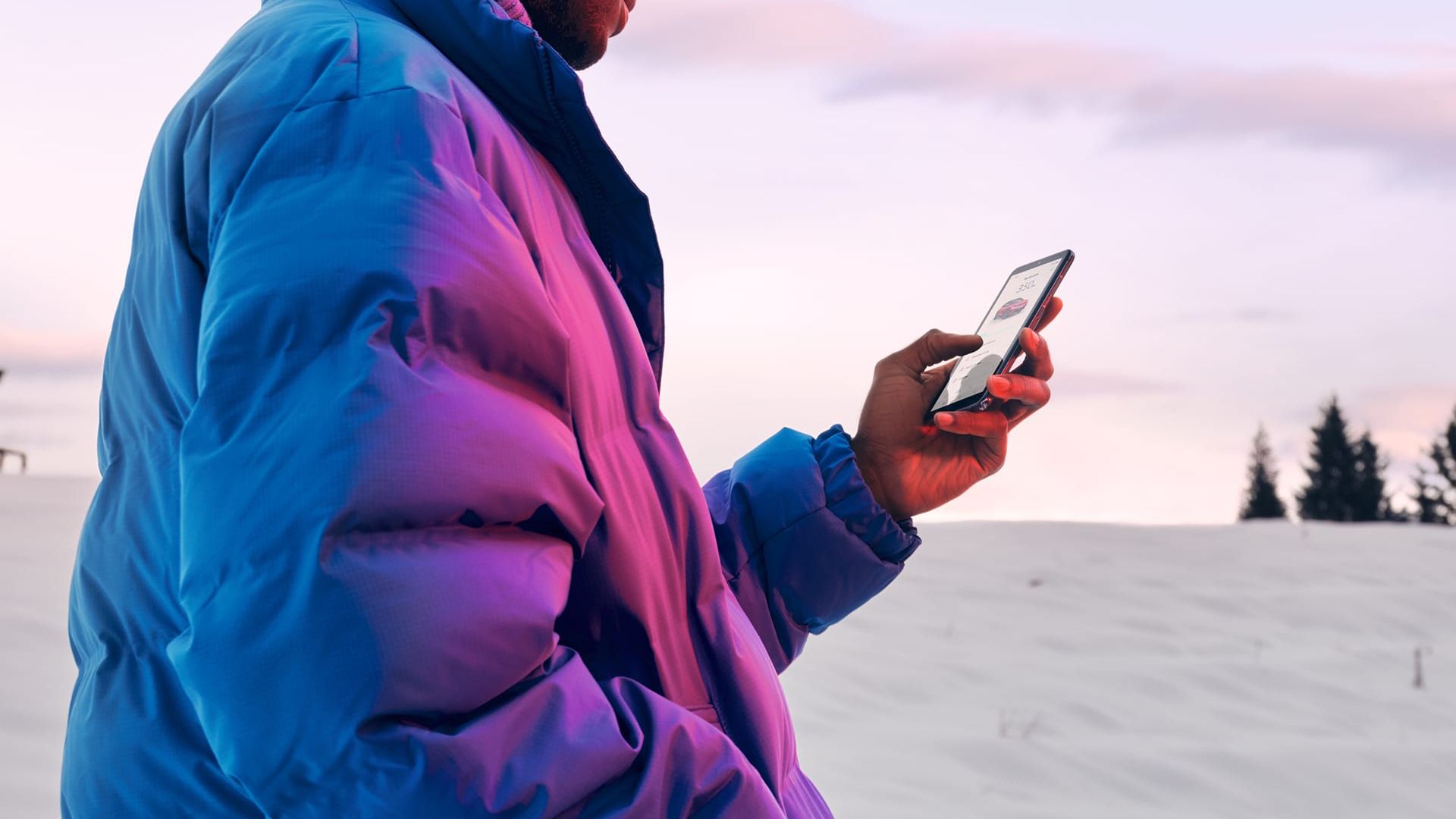 Ein Mann in Winterjacke steht in verschneiter Landschaft und schaut auf sein Smartphone.