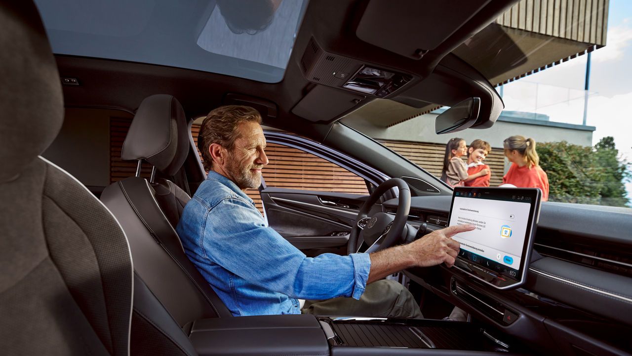 Mann sitzt in seinem Volkswagen und berührt das Display, im Hintergrund seine Familie