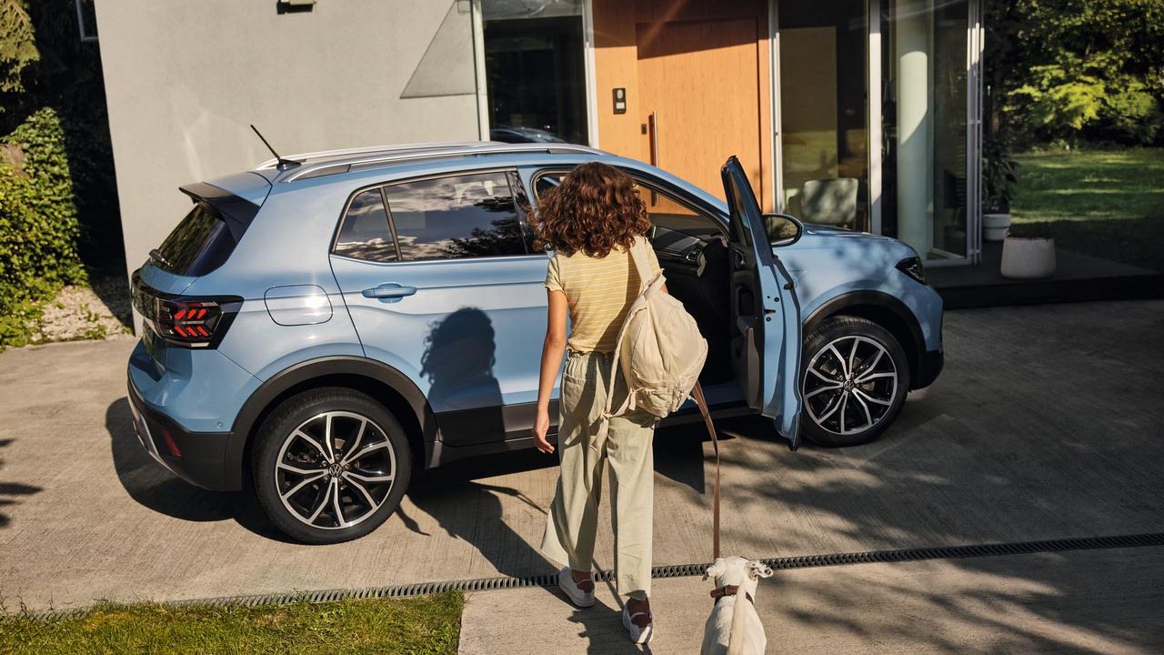 Deklica se s psom približuje modremu VW T-Crossu, ki je parkiran na dovozu. Sovoznikova vrata so odprta.