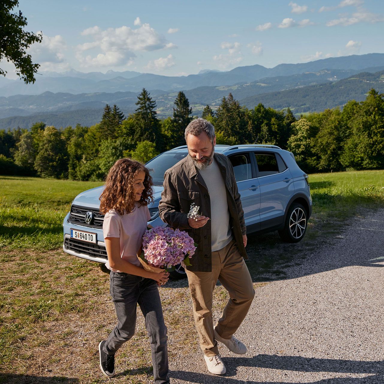 Modri VW T-Cross je parkiran na travniku v gorah. Pred njim se dekle in moški sprehajata s cvetjem v rokah.