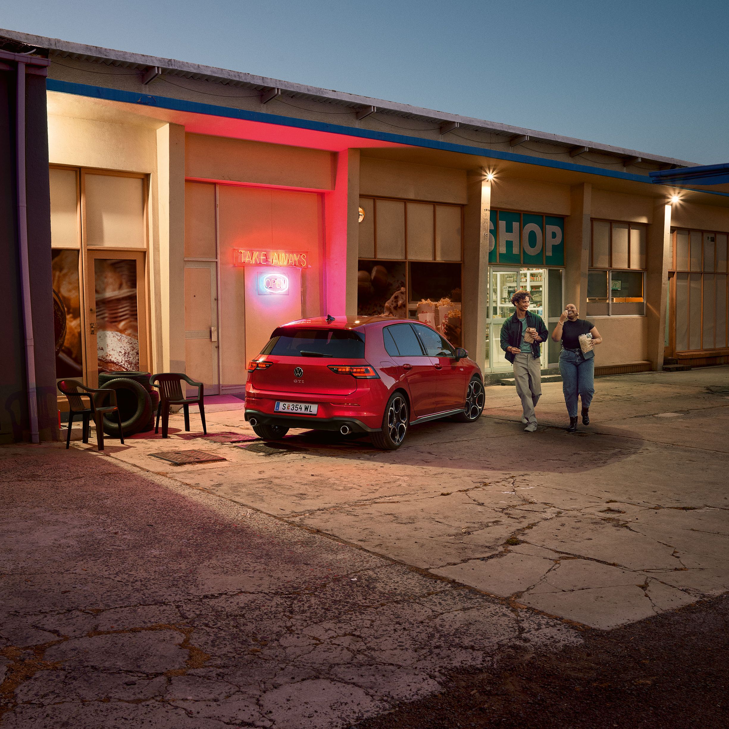 Heckansicht eines roten VW Golf GTI in abendlicher Stimmung, zwei Personen gehen neben ihm her