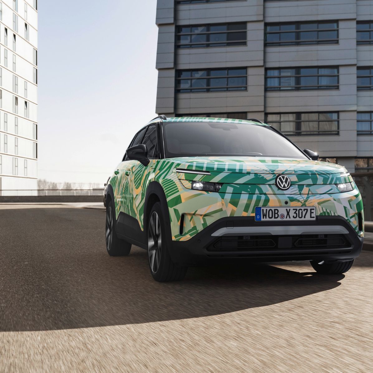 Volkswagen ID. Cross z zeleno-rumenim kamuflažnim vzorcem vozi po mestni cesti med sodobnimi večnadstropnimi stavbami. Avto je posnet iz nizkega kota spredaj levo, kar poudari njegovo višino in robustnost. 