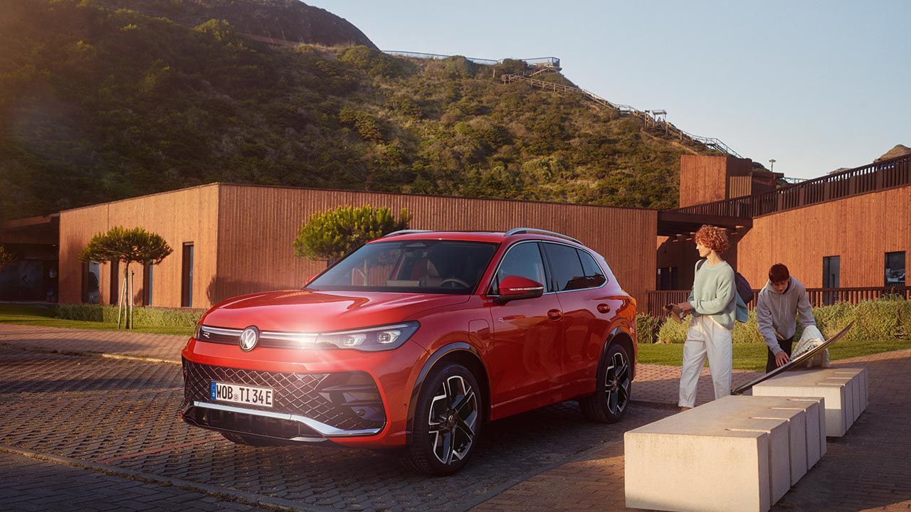 Svetlo rdeč Volkswagen Tiguan je parkiran na tlakovanem dvorišču pred sodobno zgradbo iz opeke in lesa v sončnem vremenu. V ozadju se poševno dvigajo zelene vzpetine. Na desni strani stojita dve osebi, ki se pogovarjata ob nizkem belem betonskem zidu.