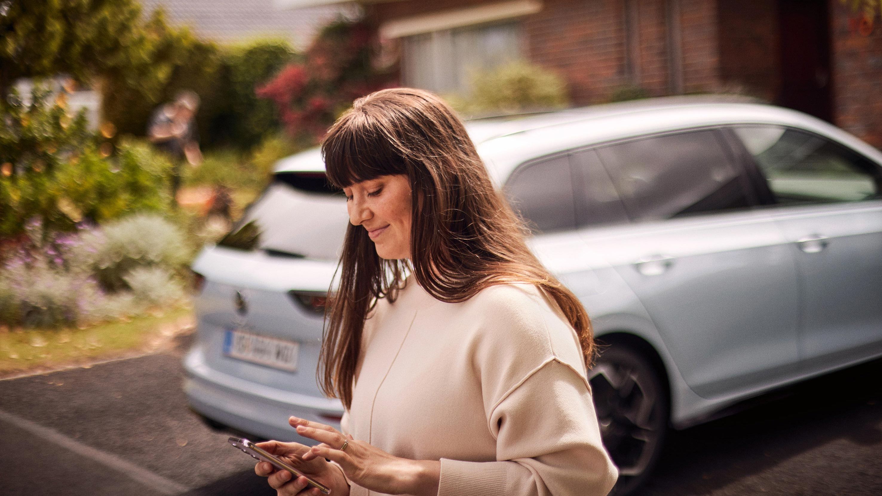 Mlajša ženska stoji za belim VW Golfom Variant in uporablja svoj pametni telefon.
