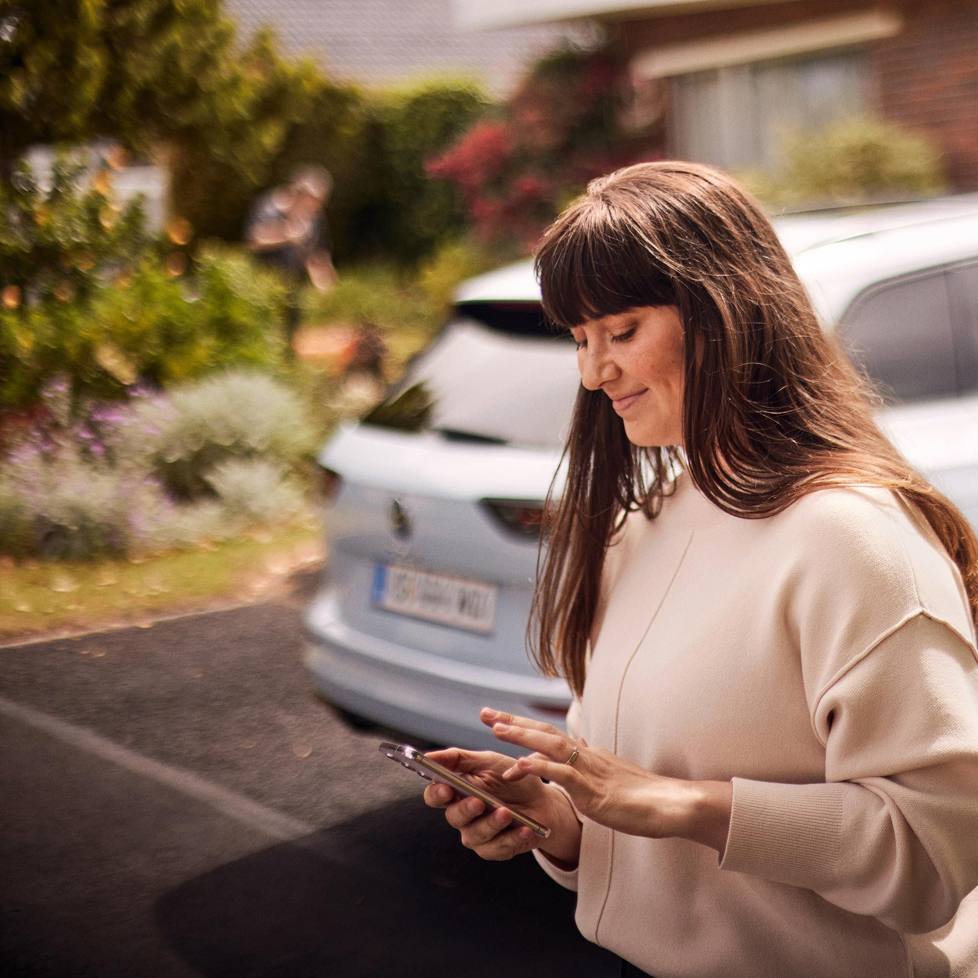 Mlajša ženska stoji za belim VW Golfom Variant in uporablja svoj pametni telefon.