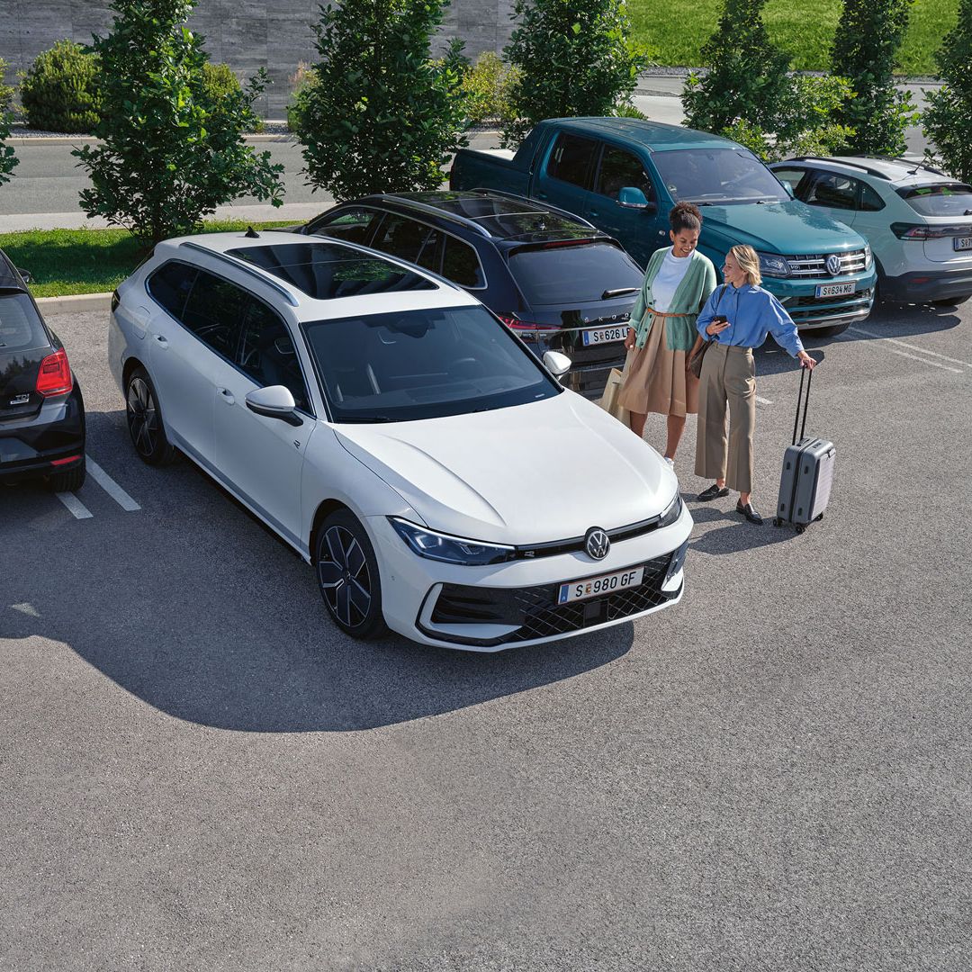 Dve osebi stojita na parkirišču poleg belega avtomobila Volkswagen. Ena oseba je oblečena v modro srajco in drži majhen kovček. Za avtomobilom je parkiranih več drugih vozil.